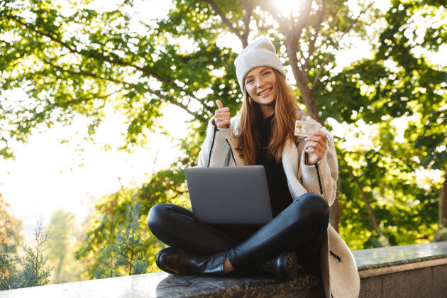 休闲快乐的年轻女子穿着秋大衣 戴着秋帽坐在户外 用笔记本电脑 展示塑料信用卡女孩使用笔记本电脑