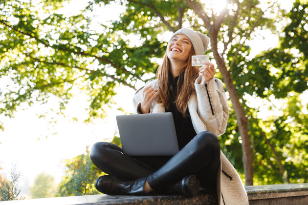 坐着快乐的年轻女子穿着秋大衣 戴着秋帽坐在户外 用笔记本电脑 展示塑料信用卡冲浪使用高加索