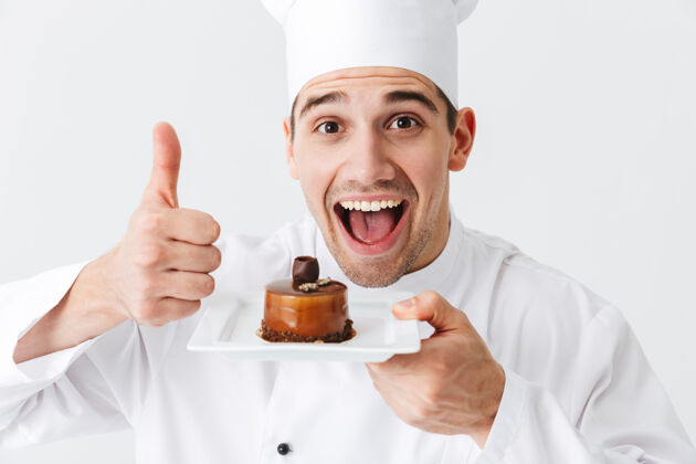 帽子开朗的厨师穿着制服 在白色的墙上展示一个盘子上的糕点烹饪美食家专业