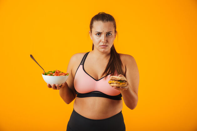 营养一个心烦意乱的超重健身妇女穿着运动服 孤立地站在黄色的墙上 手里拿着盛着沙拉和汉堡的碗午餐犹豫女孩