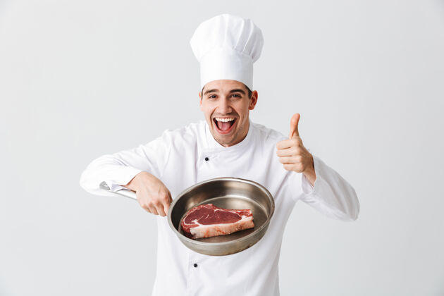 家伙开朗的厨师身着制服 在白墙上展示一个生牛排的煎锅 竖起大拇指平底锅食谱原料