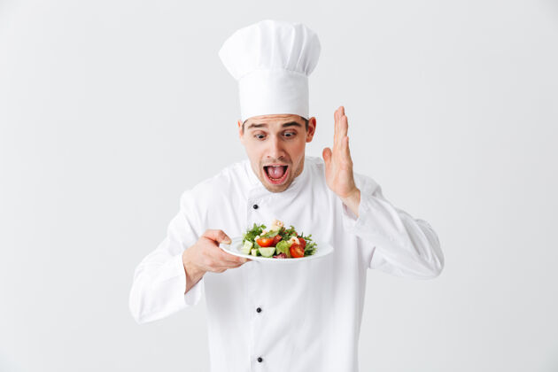 烹饪兴奋的男厨师穿着制服 把新鲜的蔬菜沙拉放在隔离在白色墙上的盘子上举行制服膳食
