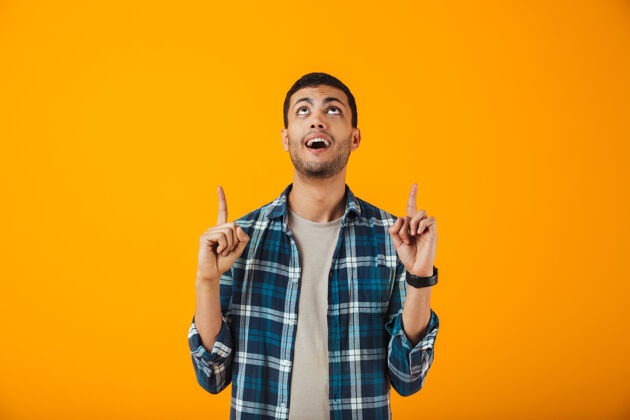 白种人一个穿着格子衬衫的快乐的年轻人孤零零地站在橙色的墙上 用手指着复制空间年轻人手势