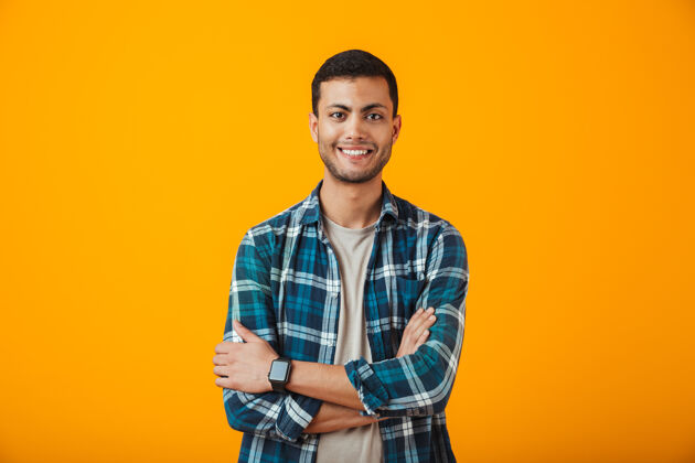 男性一个穿着格子衬衫的快乐的年轻人孤零零地站在橙色的墙上 双臂交叉着手看朋友