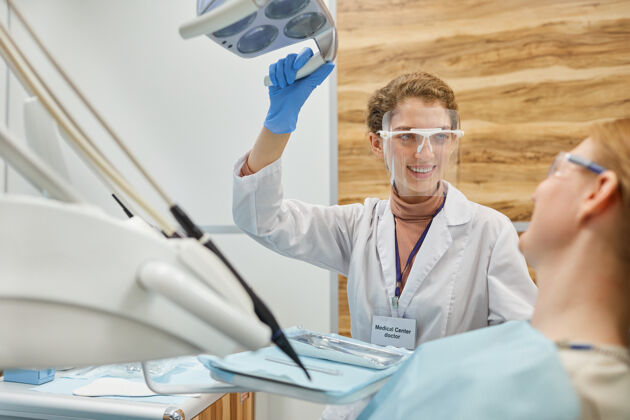 实验服戴着护目镜的年轻牙医在诊所的牙科手术中对病人微笑室内眼镜工作
