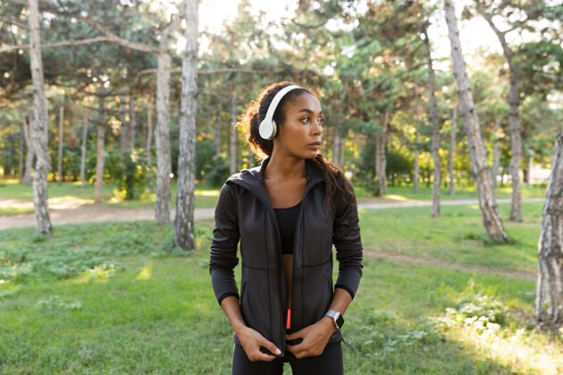 吸引力20多岁的黑发女子穿着黑色运动服 戴着耳机 穿过绿色公园散步保健耳机