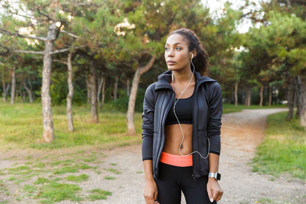 跑步20多岁的美女穿着黑色运动服 戴着耳机 穿过绿色公园运动健身运动