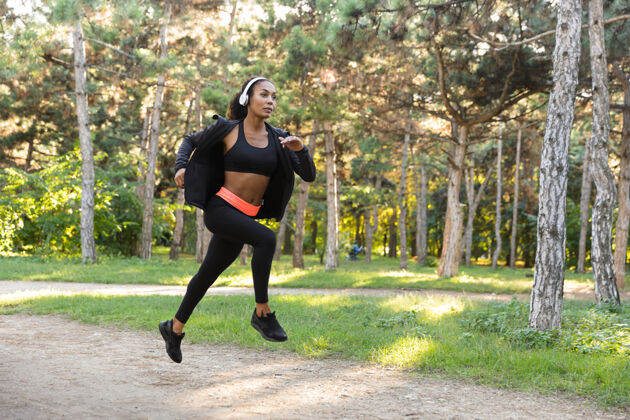 锻炼一位20多岁的女运动员穿着黑色运动服 戴着耳机 在绿色公园里跑步身体女运动员深色头发