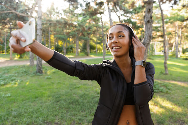 跑步一张20多岁的微笑女子的肖像 她穿着黑色运动服 戴着耳机 在绿色公园散步时用手机自拍自然女性女运动员