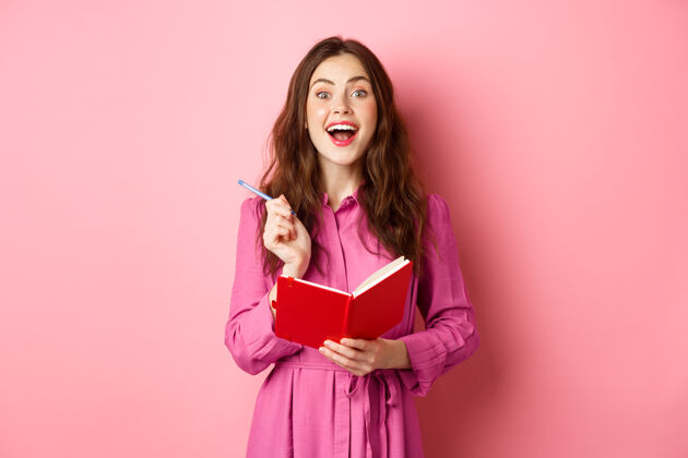 购物兴奋的年轻女子有很好的想法 把她的想法写在计划者 拿着笔记本日记 微笑着惊讶 站在粉红色的墙上笔记本快乐女孩