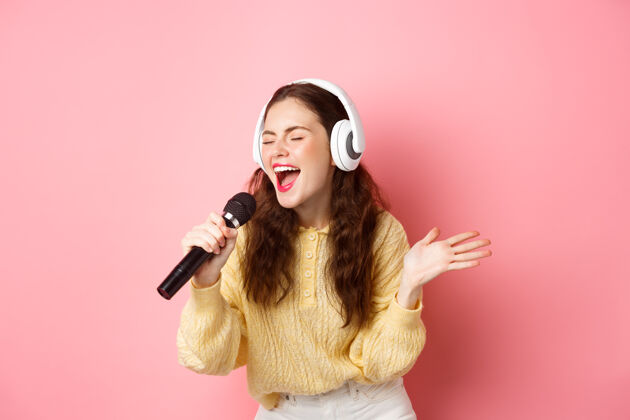 时尚年轻女子唱卡拉OK 戴着耳机 拿着麦克风 站在粉红色的墙上卡拉OK水平微笑
