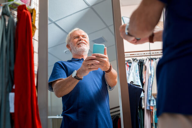欢乐新画面快乐的白发男人一边用手机一边对着镜子自拍一边欣赏自己的倒影时尚老年人老年人