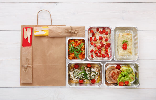 餐具健康食品配送 日常膳食和零食营养 蔬菜 肉类和水果在铝箔盒和牛皮纸包装俯视图 平放在白色木材与复印空间包装外卖晚餐