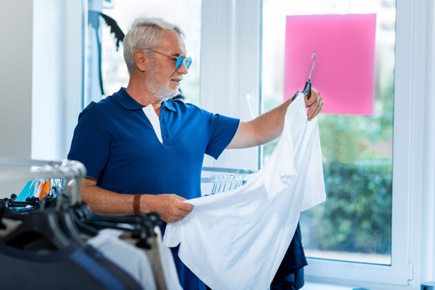 年龄逛街时尚正面的灰头发男人站在商店里 一边好奇地选择新的白色t恤消费太阳镜老年人