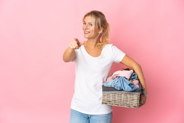 年轻一个年轻的俄罗斯女人拿着一个独立于粉色背景的衣篮 脸上带着幸福的表情指着前面洗衣服俄罗斯