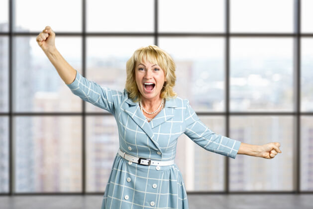 握紧兴奋的成熟女人紧握着拳头快乐的女人紧握着拳头在办公室窗户的不同侧面兴奋人类胜利的表情人专业成熟