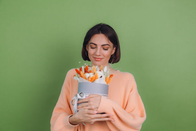 女人年轻女子穿着休闲桃色毛衣隔离在绿橄榄墙上手持橙白色花盒组成的棉花 吉普赛拉小麦和拉古鲁斯作为礼物开心惊喜魅力花蝴蝶结