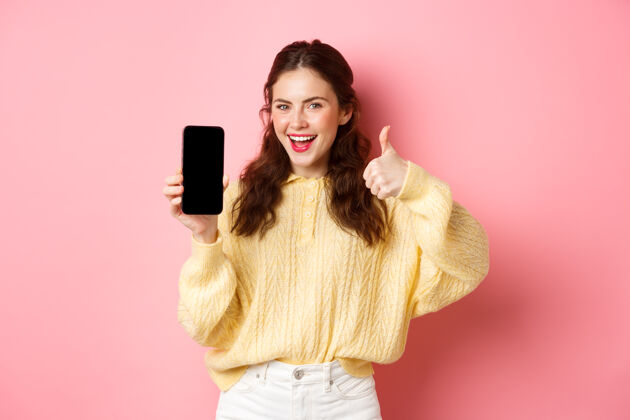 竖起大拇指微笑着 满意的女人竖起大拇指 空着智能手机屏幕 推荐应用程序或网站 站在粉色的墙上应用程序崇拜候选人