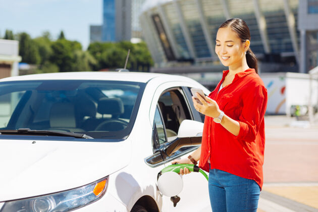 车辆现代自信的年轻女子拿着加油嘴看着她的智能手机城市人环境