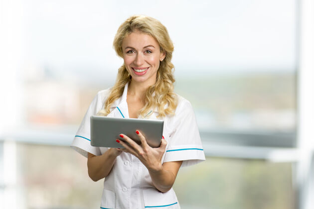诊所快乐的年轻护士拿着电脑平板微笑的年轻女孩穿着白色制服拿着数码平板 看着相机 诊所的窗户女性个人医学