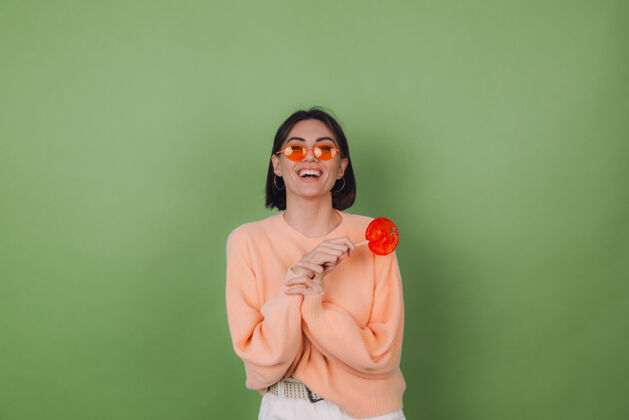 女性年轻时尚的女士穿着休闲的桃色毛衣 戴着橙色的眼镜 隔离在绿色橄榄色的墙上 带着橙色棒棒糖的正面微笑复制空间糖果积极甜点