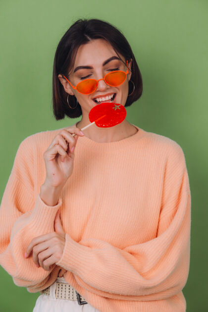 时尚年轻时尚的女士穿着休闲的桃色毛衣 戴着橙色的眼镜 隔离在绿色橄榄色的墙上 带着橙色棒棒糖的正面微笑复制空间青少年肖像甜点