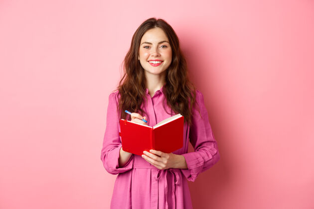 兴奋开朗的女孩微笑着写下笔记 拿着她的计划者 计划表或杂货清单 拿着日记本站在粉红色的墙上女孩手势策划