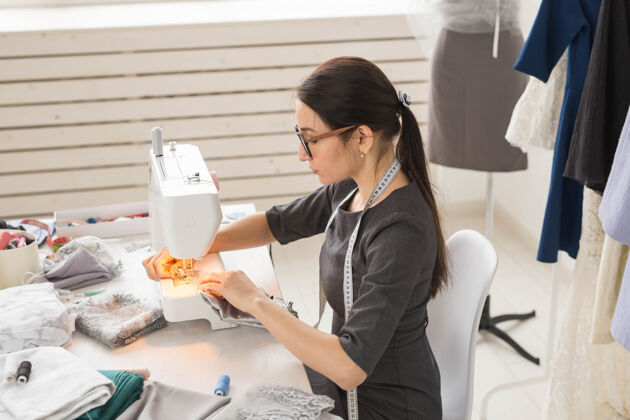 缝纫人 裁缝和时尚概念-侧视图肖像的时装设计师在眼镜使用缝纫机在车间女性时尚工作场所
