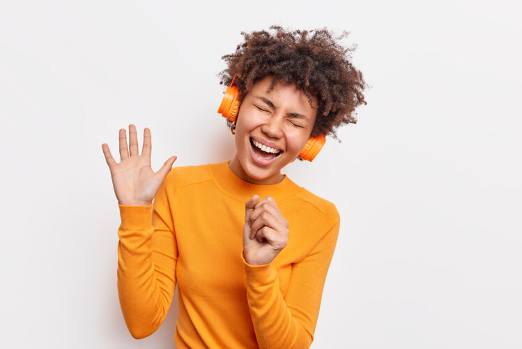 音乐喜出望外的非洲裔美国妇女一直掌心高举有无忧无虑的表情唱歌在耳机里听音乐穿着休闲的橙色套头衫隔着白墙有趣的娱乐表达欢呼舞蹈