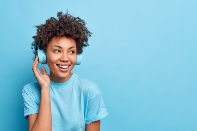 女人这张照片拍摄的是一个穿着休闲服 留着非洲裔头发的少女 她通过无线耳机收听播放列表中的音乐 这张照片被隔离在蓝色墙壁上 为您的广告复制空间可穿戴穿女孩