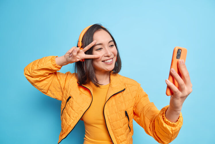 自拍快乐的亚洲女人微笑愉快地做和平手势在眼睛上自拍现代智能手机通过立体声无线耳机听音乐隔着蓝色的墙生活方式技术学生欢呼快乐