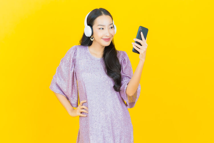 音频年轻的亚洲女人 戴着耳机和智能手机在黄色屏幕上听音乐人物手机乐趣