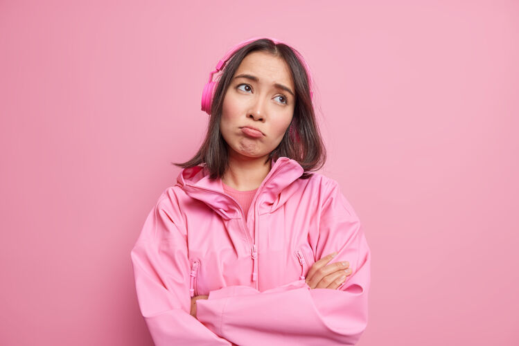 关心失意失望的亚洲女人愁眉苦脸 双臂合十 心无旁骛 通过无线耳机听音乐 穿着隔离在粉色墙上的夹克问题女人女性