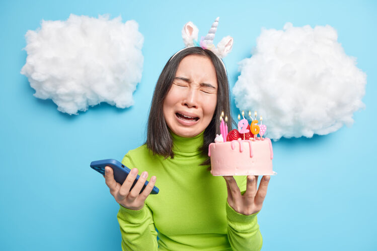 忧郁女人哭着拿着智能手机和生日蛋糕心烦意乱没有人祝贺她感到孤独沮丧戴着独角兽头带绿色圆领高领毛衣手机场合