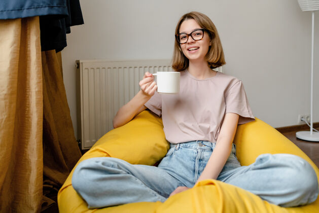 女人坐在公寓里微笑的年轻女子 穿着蓝色牛仔裤 粉色T恤 戴着眼镜 手里拿着一个杯子杯子眼镜包椅
