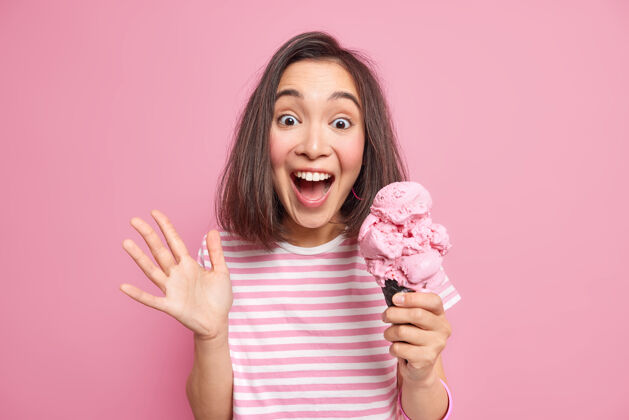 反应情绪化的黑发女人大声地把手掌举起来吃美味的冰淇淋对一些意想不到的事情做出反应令人惊讶的快乐表情穿着休闲条纹t恤隔离在粉红色的墙上条纹可口食物