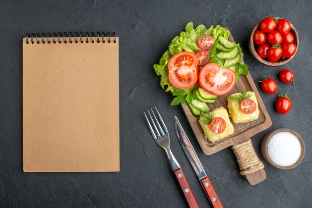 橡皮上图是切好的新鲜西红柿和黄瓜 奶酪放在木板上 餐具放在黑色表面的盐笔记本上食物刀健康