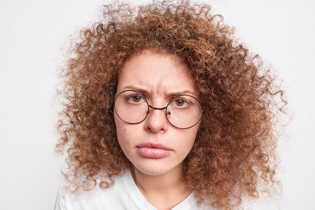欧洲人愤怒恼怒的欧洲年轻女人头上的头发卷曲浓密 戴着圆眼镜隔着白墙 看起来很不开心人类的表情和反应概念表达反应时尚