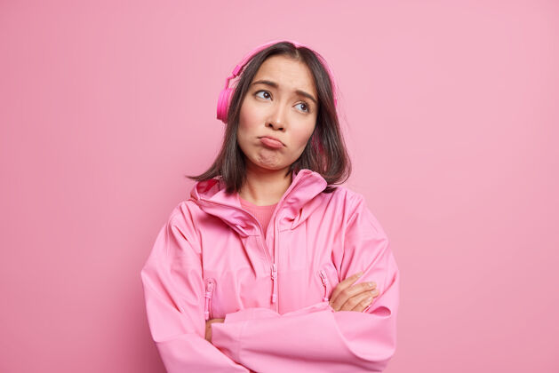 关心失意失望的亚洲女人愁眉苦脸 双臂合十 心无旁骛 通过无线耳机听音乐 穿着隔离在粉色墙上的夹克问题女人女性
