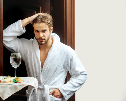 放松酒店里的男人年轻英俊的男人穿着白色毛巾浴衣在酒店附近的人手里拿着服务员的食物和托盘上的水杯公司男性工作