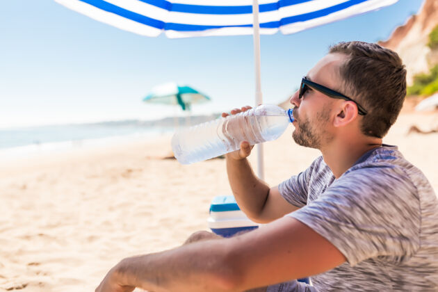 躺椅在绿色太阳伞下的年轻人在沙滩上喝水假日口渴海湾