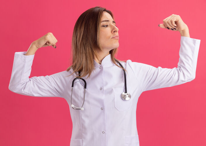 穿着自信地看着身边穿着医用长袍的年轻女医生 听诊器显示出强烈的手势 隔离在粉红色的墙上年轻壁板强壮