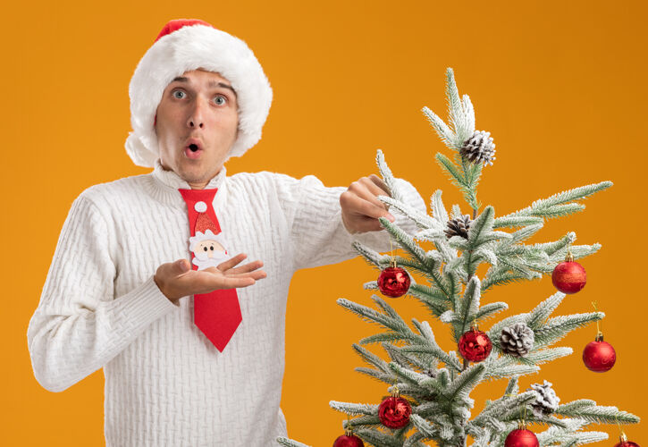 小伙子令人印象深刻的年轻帅哥戴着圣诞帽和圣诞老人领带站在圣诞树旁装饰圣诞球装饰看起来空手孤立在橙色的墙上近空帽子