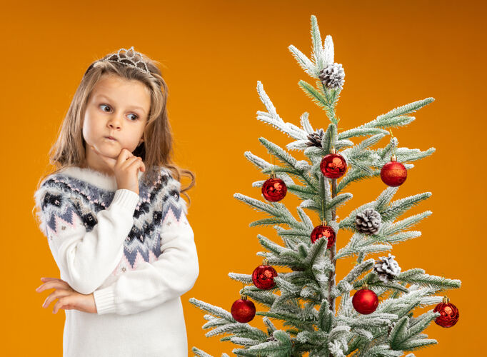手想着站在圣诞树旁戴着头饰脖子上戴着花环的小女孩把手放在下巴下面孤立在橙色的墙上附近穿头饰