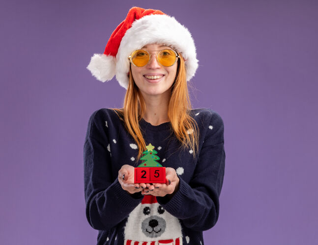 帽子微笑的年轻漂亮女孩穿着圣诞毛衣戴着圣诞帽戴着眼镜拿着圣诞玩具孤立在紫色的墙上眼镜年轻人女孩