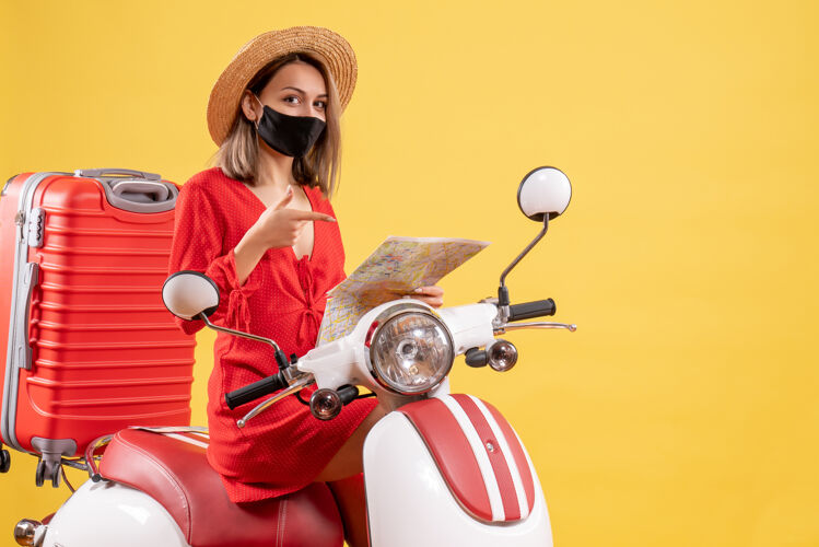 滑板车前视图：骑着轻便摩托车的穿着红裙子的年轻女士指着手中的地图旅行面罩人