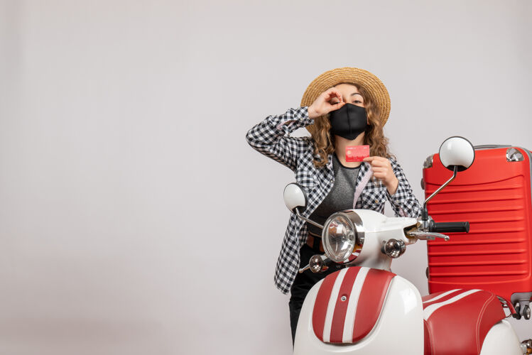 旅游者正面图：戴着黑色面具的年轻女孩手持制票望远镜站在红色轻便摩托车旁望远镜面具车辆
