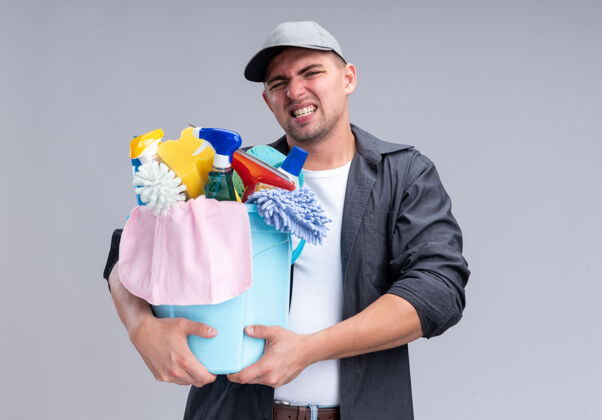 帅哥紧张的年轻帅气的保洁员穿着t恤 戴着帽子 手里拿着一桶洁具隔离在白色的墙上衣服紧张水桶