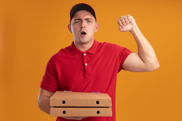 脸快乐的年轻送货员穿着制服 戴着帽子 拿着披萨盒 在橙色的墙上显示“是”的手势盒子公民人