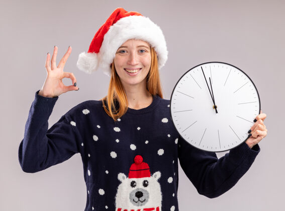 帽子微笑着的年轻漂亮女孩戴着圣诞帽拿着挂钟在白色的墙上显示出良好的姿态隔离年轻墙壁好的